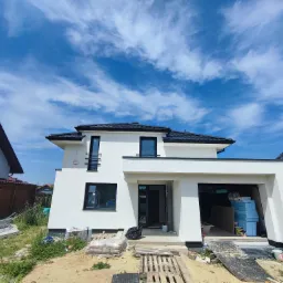 Nowoczesny, biały dom jednorodzinny w trakcie ocieplania elewacji, widoczne materiały izolacyjne w garażu i na podjeździe, błękitne niebo z chmurami w tle.