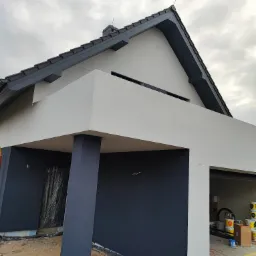 Nowoczesny dom z szarą elewacją i ciemnoszarą stolarką okienną, świeżo po malowaniu, widoczne puszki z farbą w garażu, pochmurne niebo w tle.