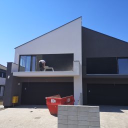 USŁUGI OGÓLNOBUDOWLANE IGOR KOŁODZIEJ BUDIGO - Dwukondygnacyjny dom w trakcie wykańczania elewacji, widoczny pracownik na rusztowaniu na balkonie, czerwony kontener na gruz przed budynkiem, nowoczesna architektura.