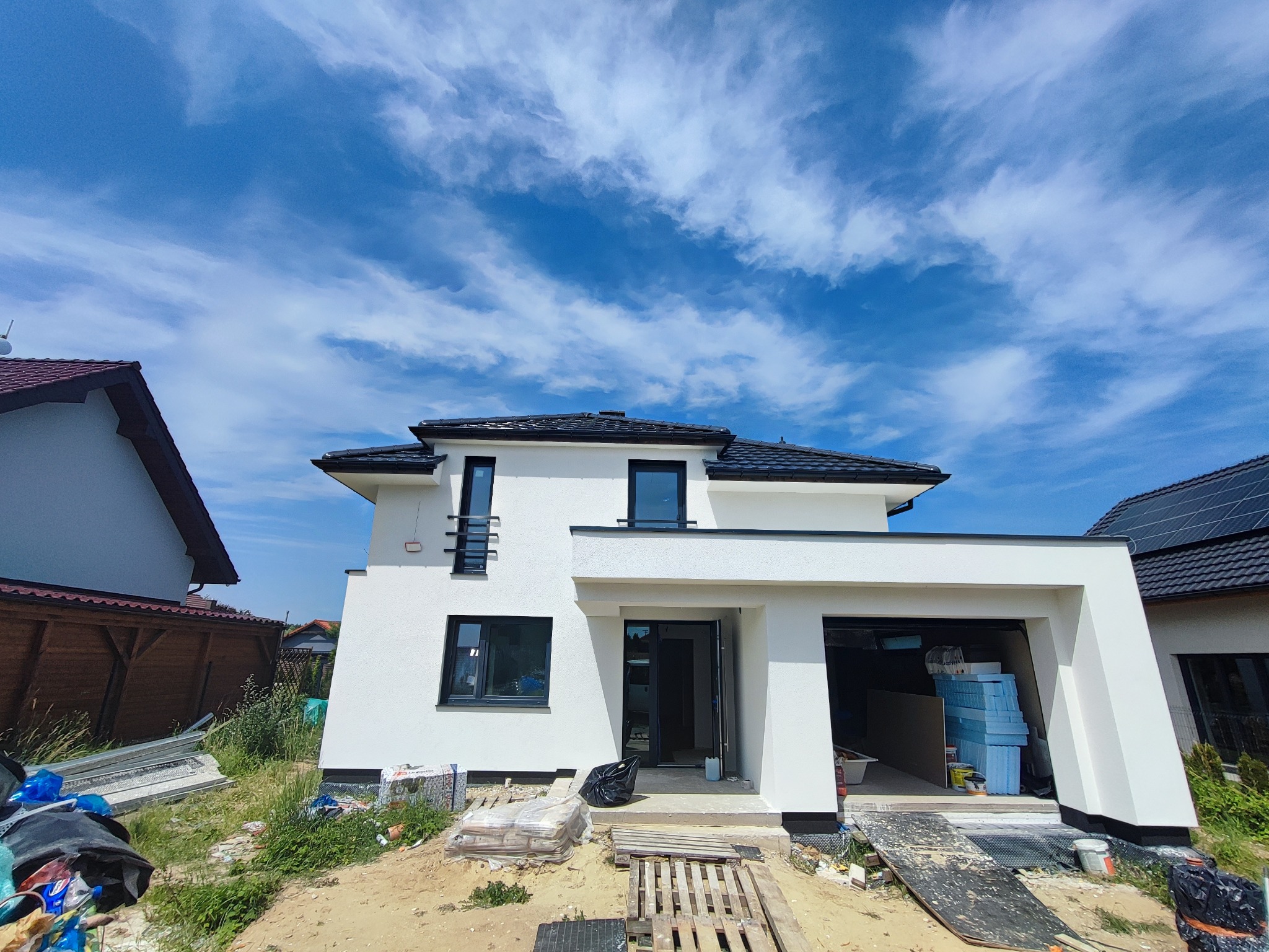 Nowoczesny, biały dom jednorodzinny w trakcie ocieplania elewacji, widoczne materiały izolacyjne w garażu i na podjeździe, błękitne niebo z chmurami w tle.