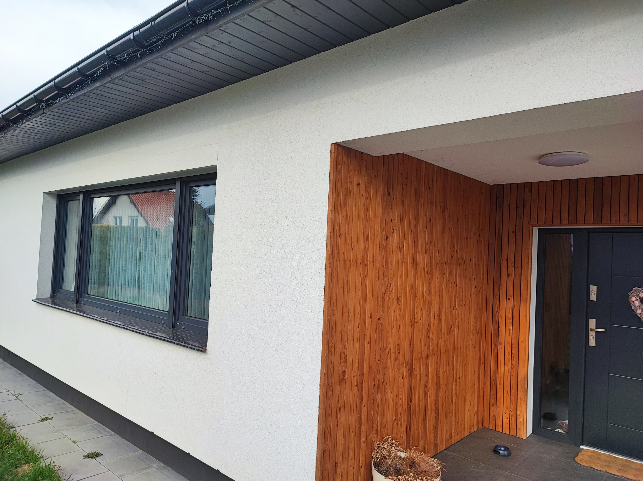 Wykończona elewacja domu jednorodzinnego z fragmentem drewnianej okładziny przy wejściu i oknem z ciemną ramą.