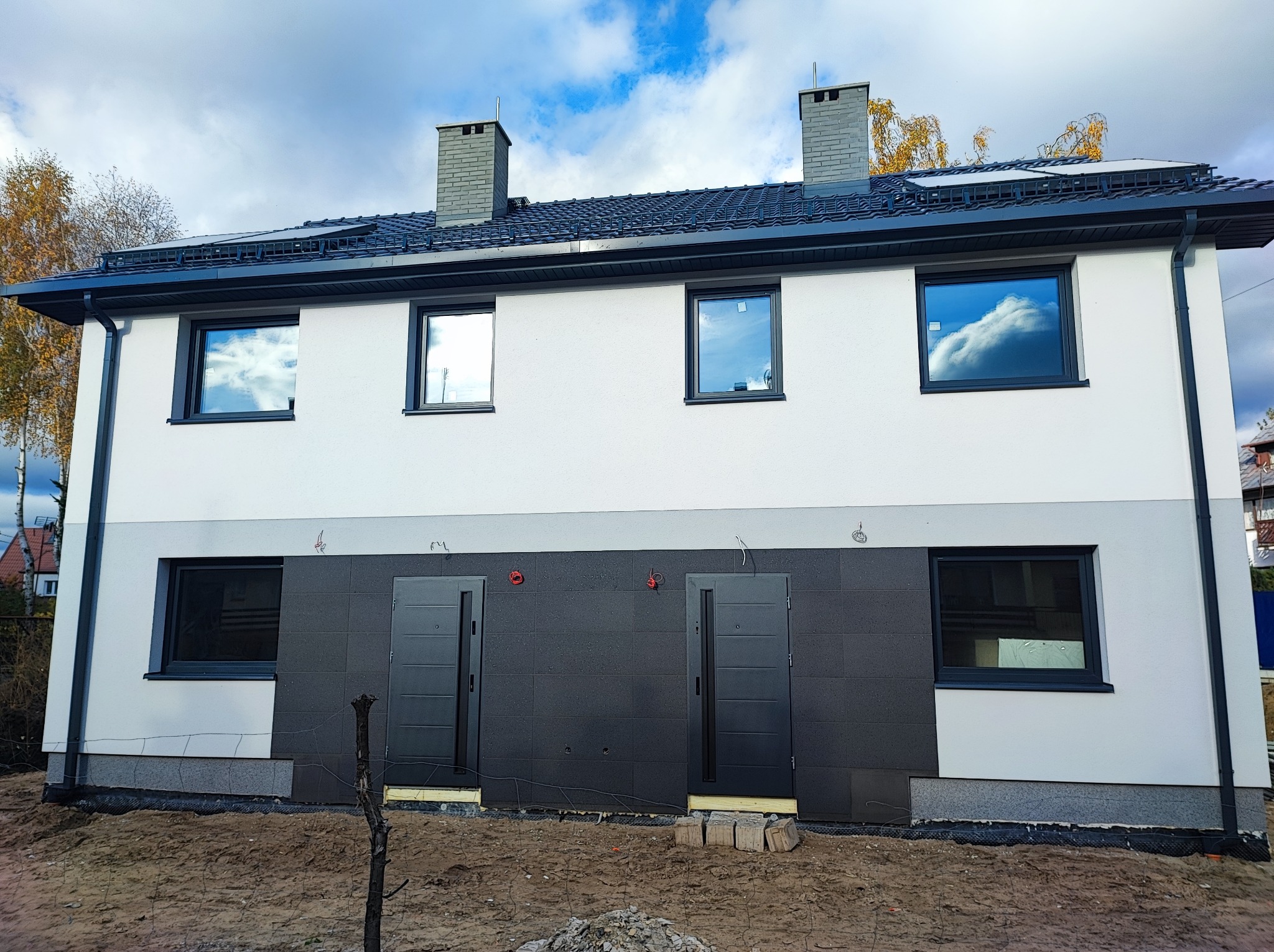 Dwukondygnacyjny dom w trakcie wykańczania elewacji, z widoczną izolacją termiczną na dolnej części ściany i świeżo pomalowaną górną częścią, okna z czarnymi ramami, dwa kominy i błękitne niebo...