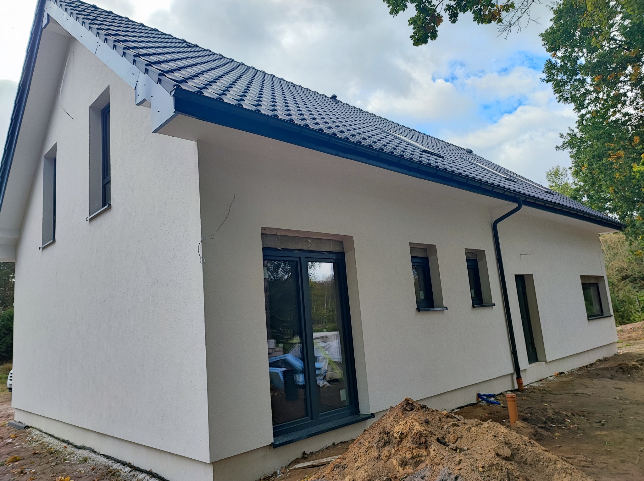 Nowoczesny dom jednorodzinny z jasną elewacją, ciemnymi oknami i dachem z czarnej dachówki, widok z zewnątrz na świeżo wykończoną ścianę i przygotowany teren.