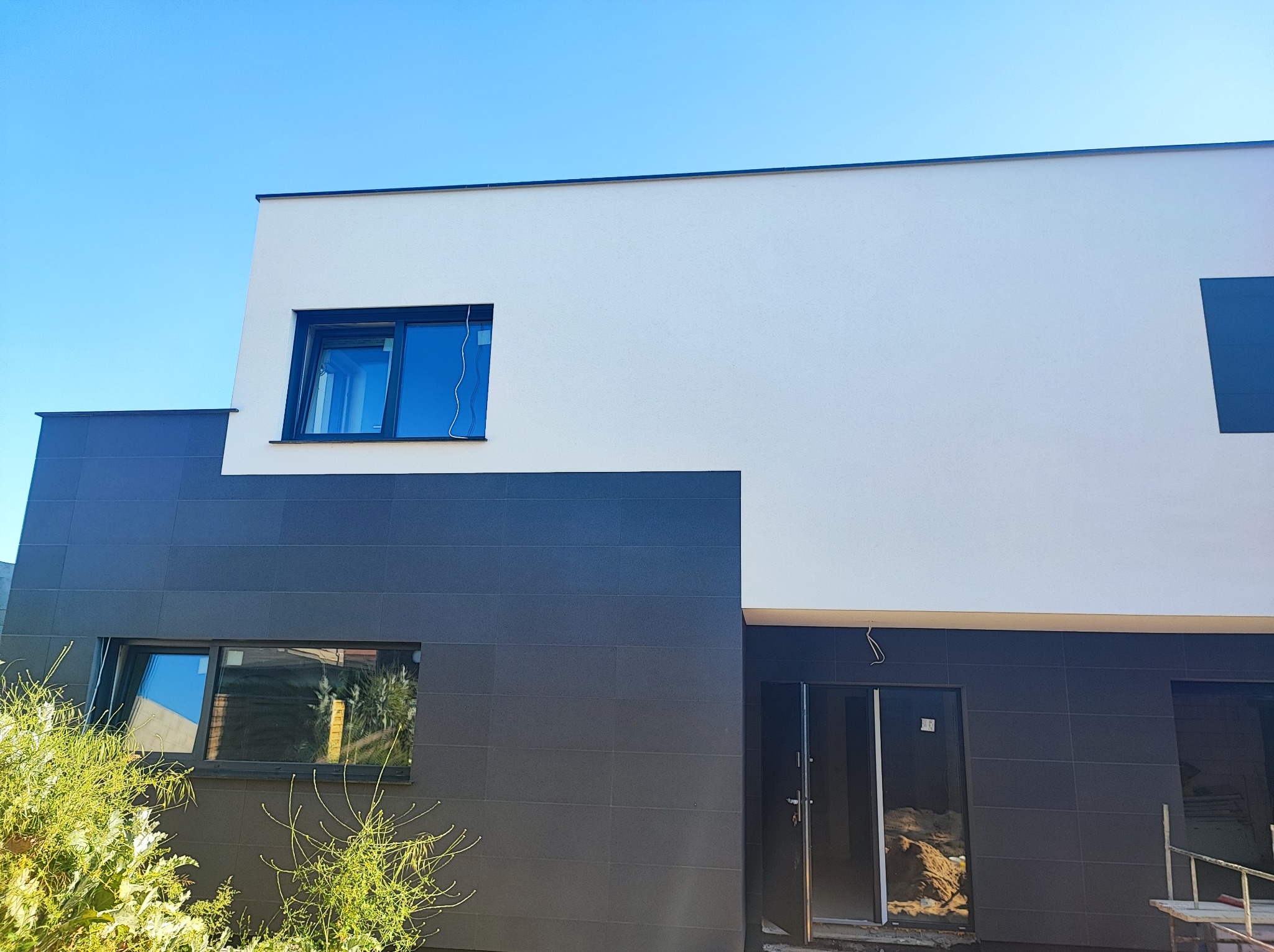 Dwukolorowa elewacja nowoczesnego domu z czarnymi oknami, w tle błękitne niebo, widoczne rośliny w dolnej części kadru.