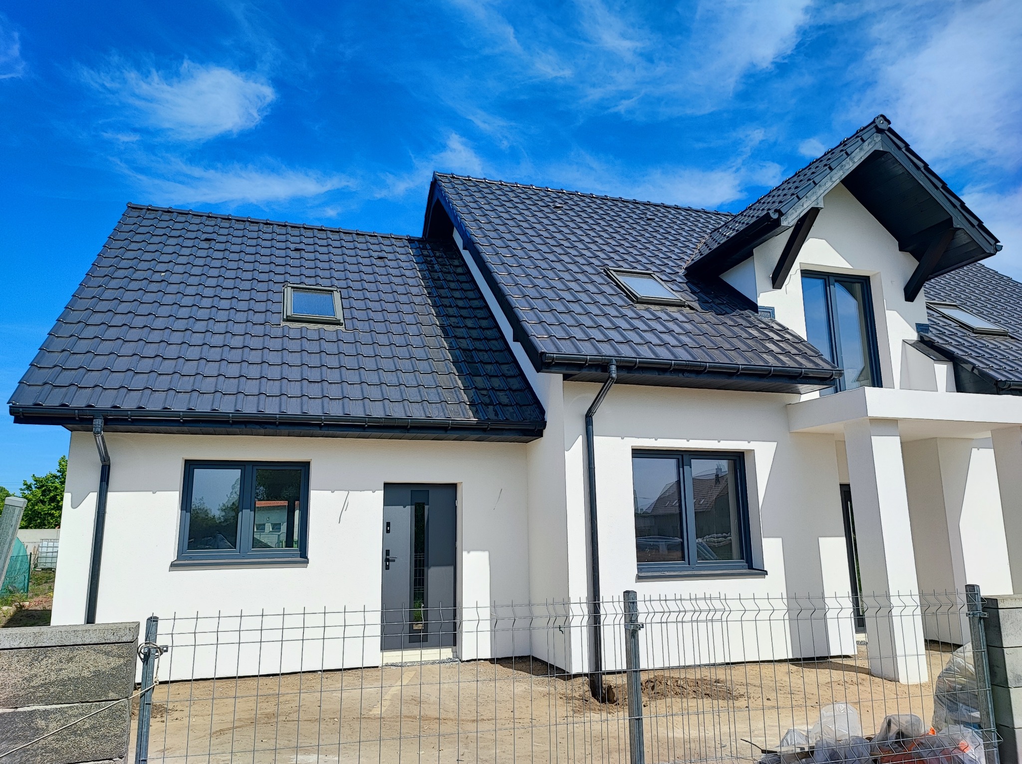 Nowoczesny dom jednorodzinny z białą elewacją, grafitową stolarką okienną i dachówką, uchwycony w słoneczny dzień z widocznymi elementami ogrodzenia.