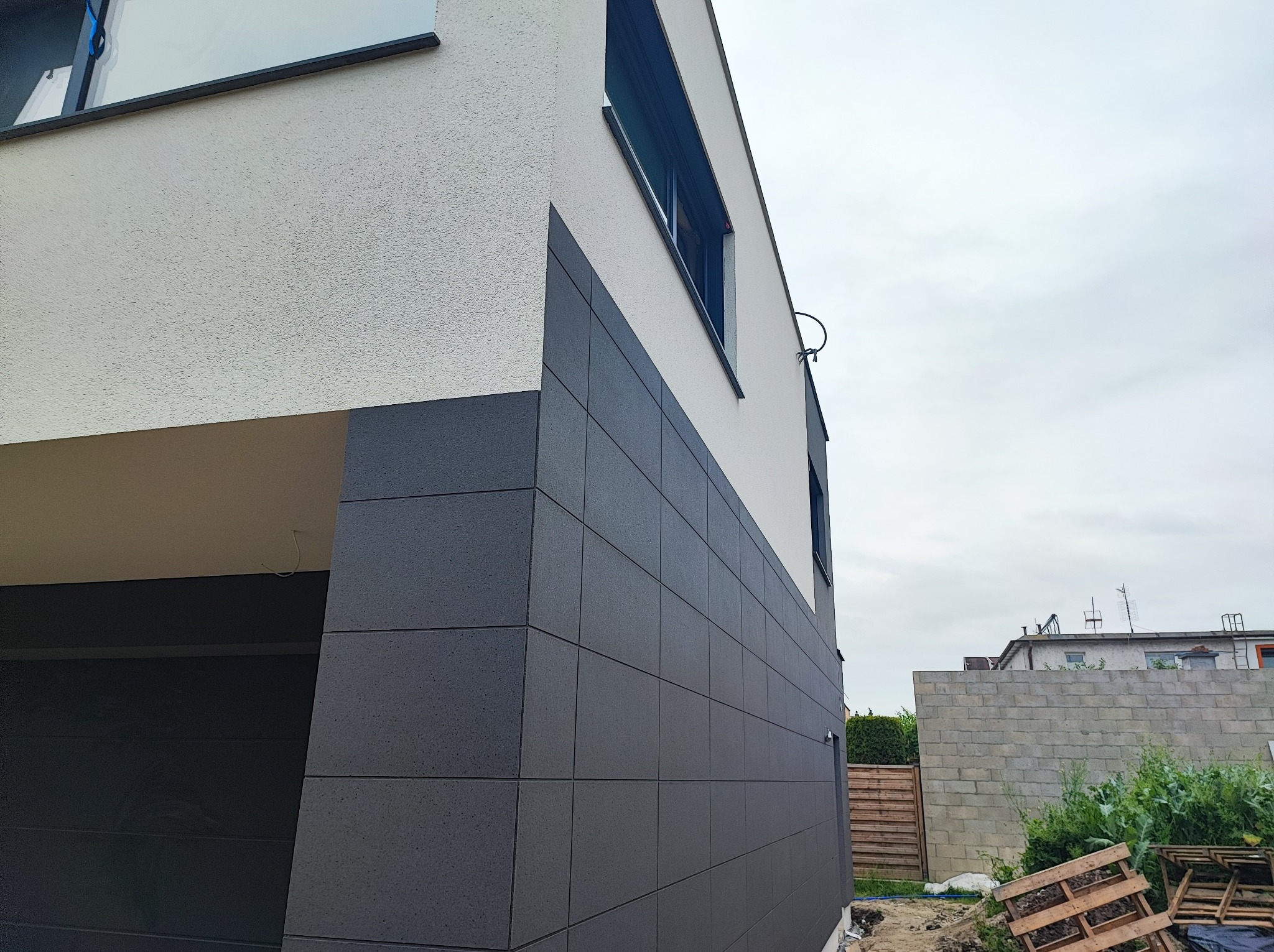 Nowoczesna elewacja domu z szarymi płytami elewacyjnymi i białym tynkiem strukturalnym, widok z boku na narożnik budynku, zarys okna z roletą.