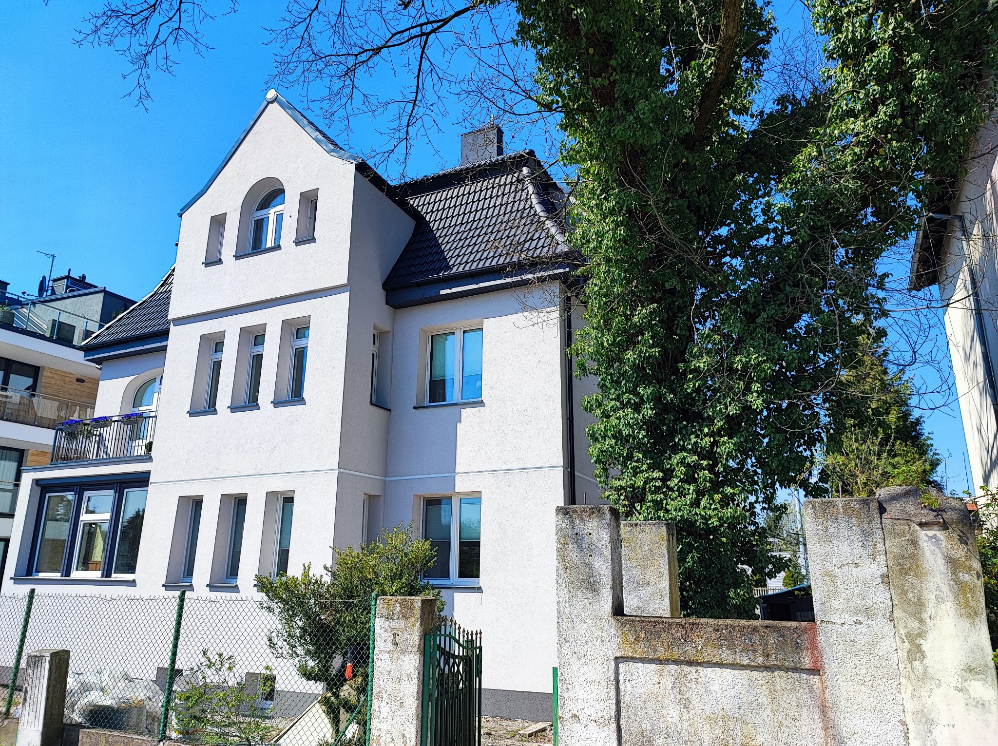 Dwupiętrowy dom z białą elewacją, czarnym dachem i charakterystycznymi oknami, częściowo zasłonięty przez drzewo i ogrodzenie.