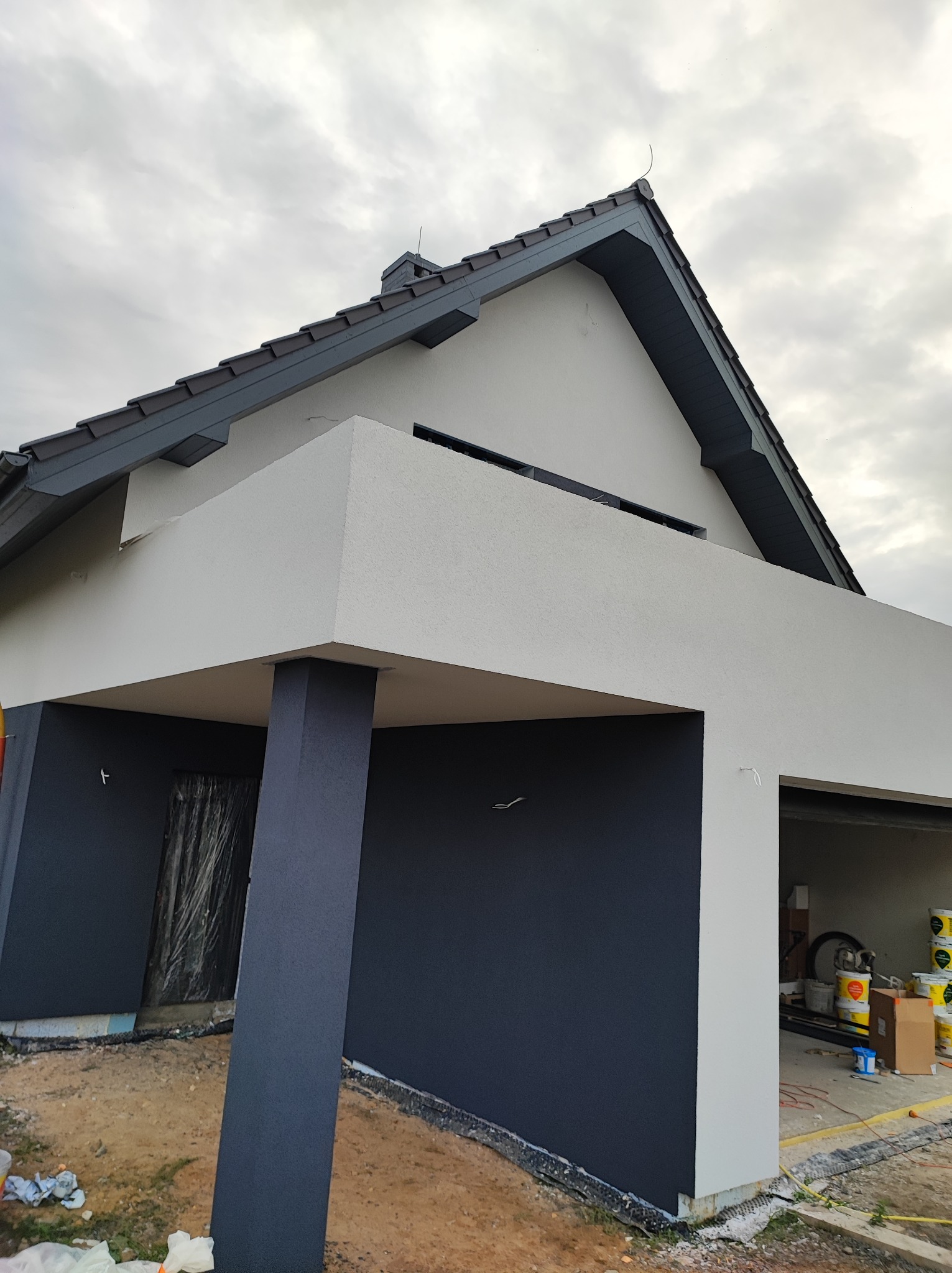 Nowoczesny dom z szarą elewacją i ciemnoszarą stolarką okienną, świeżo po malowaniu, widoczne puszki z farbą w garażu, pochmurne niebo w tle.