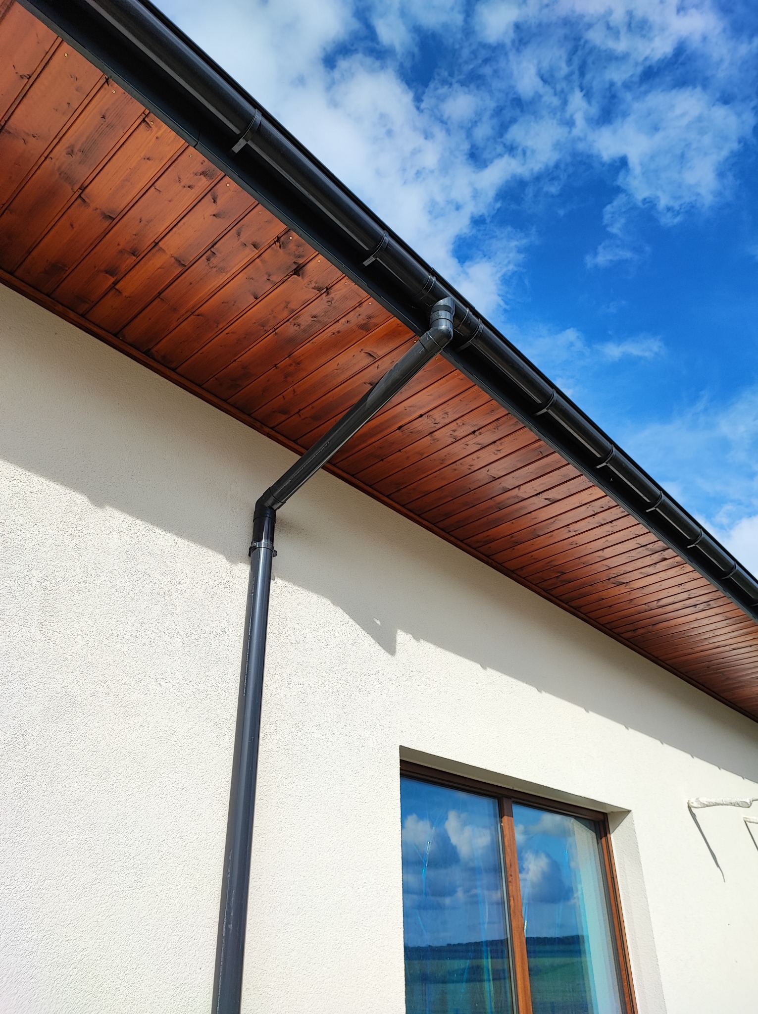 Fragment elewacji domu z drewnianą podbitką i systemem rynnowym w kolorze antracytowym, na tle błękitnego nieba z chmurami.