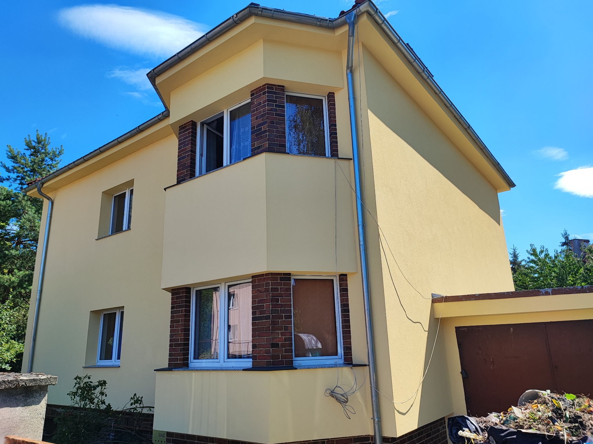 Żółty dom jednorodzinny z ceglanymi elementami dekoracyjnymi wokół okien, świeżo po malowaniu, widoczne ślady prac na podjeździe.