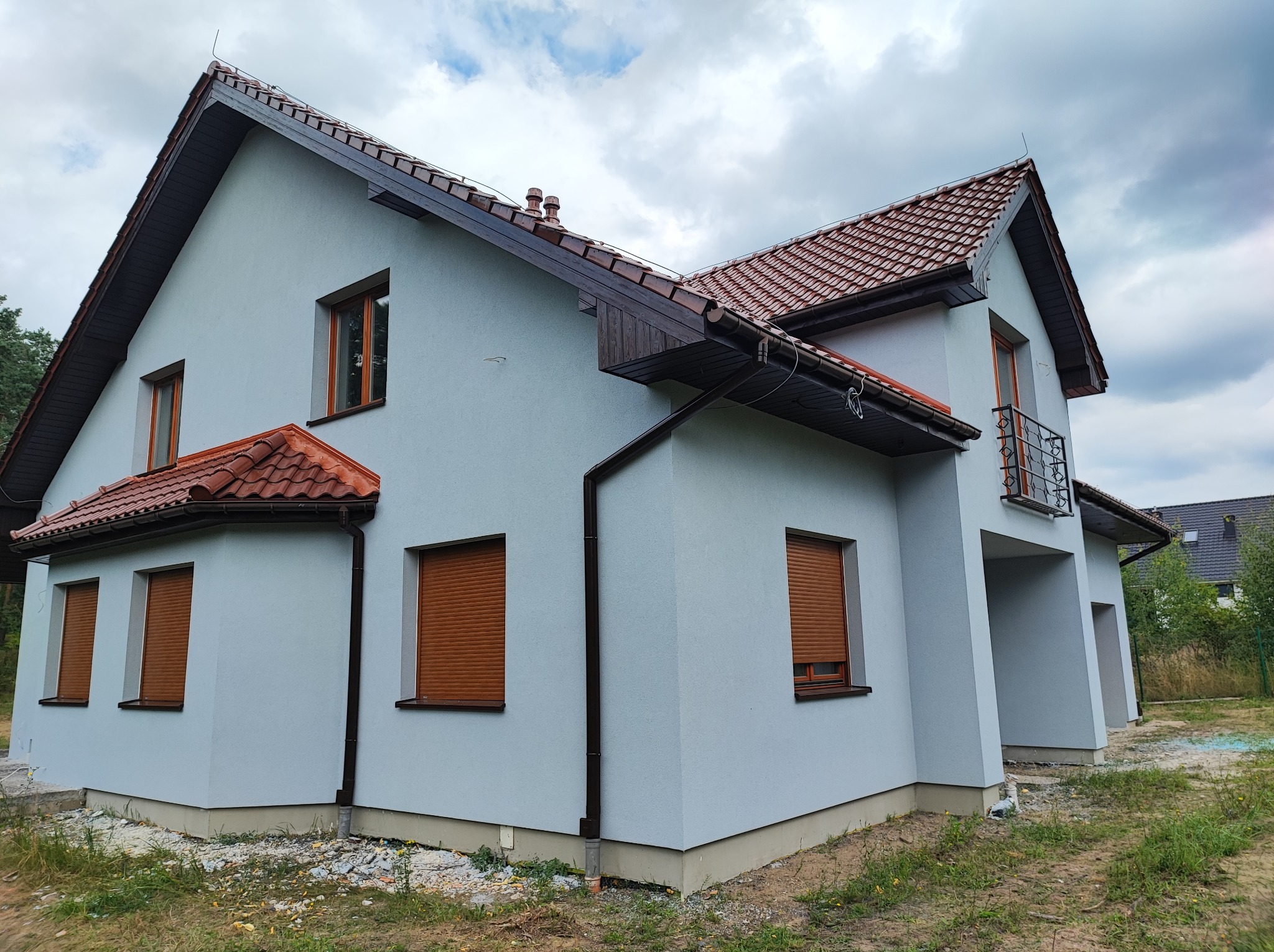 Dwukondygnacyjny dom jednorodzinny z szarą elewacją, brązową dachówką i roletami zewnętrznymi w oknach, widoczny z perspektywy narożnej, na tle pochmurnego nieba.