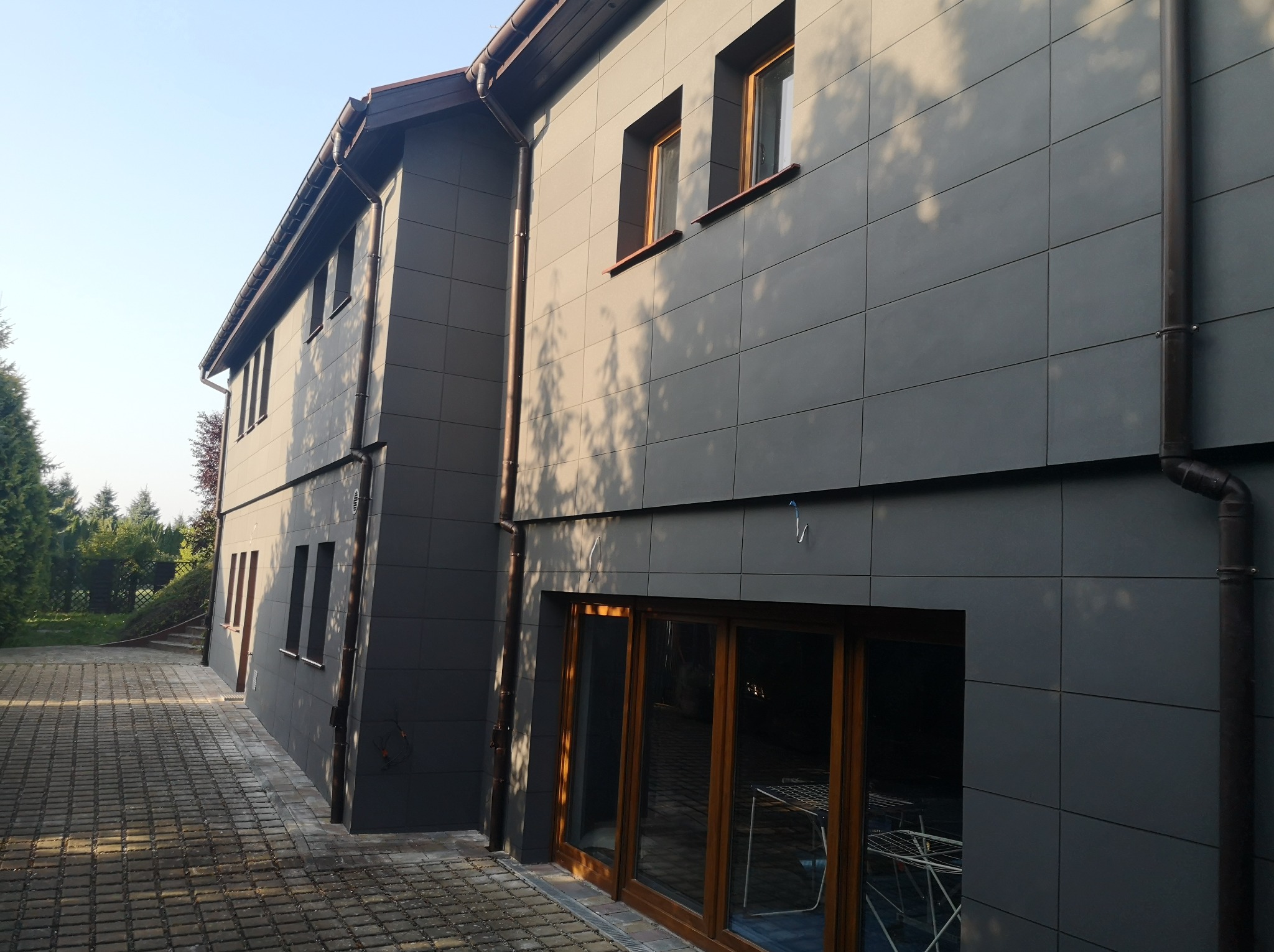 Nowoczesny budynek z szarą elewacją panelową, drewniane okna i drzwi tarasowe, widoczny fragment brukowanego podjazdu i drzewa w tle, cień drzew na ścianie.