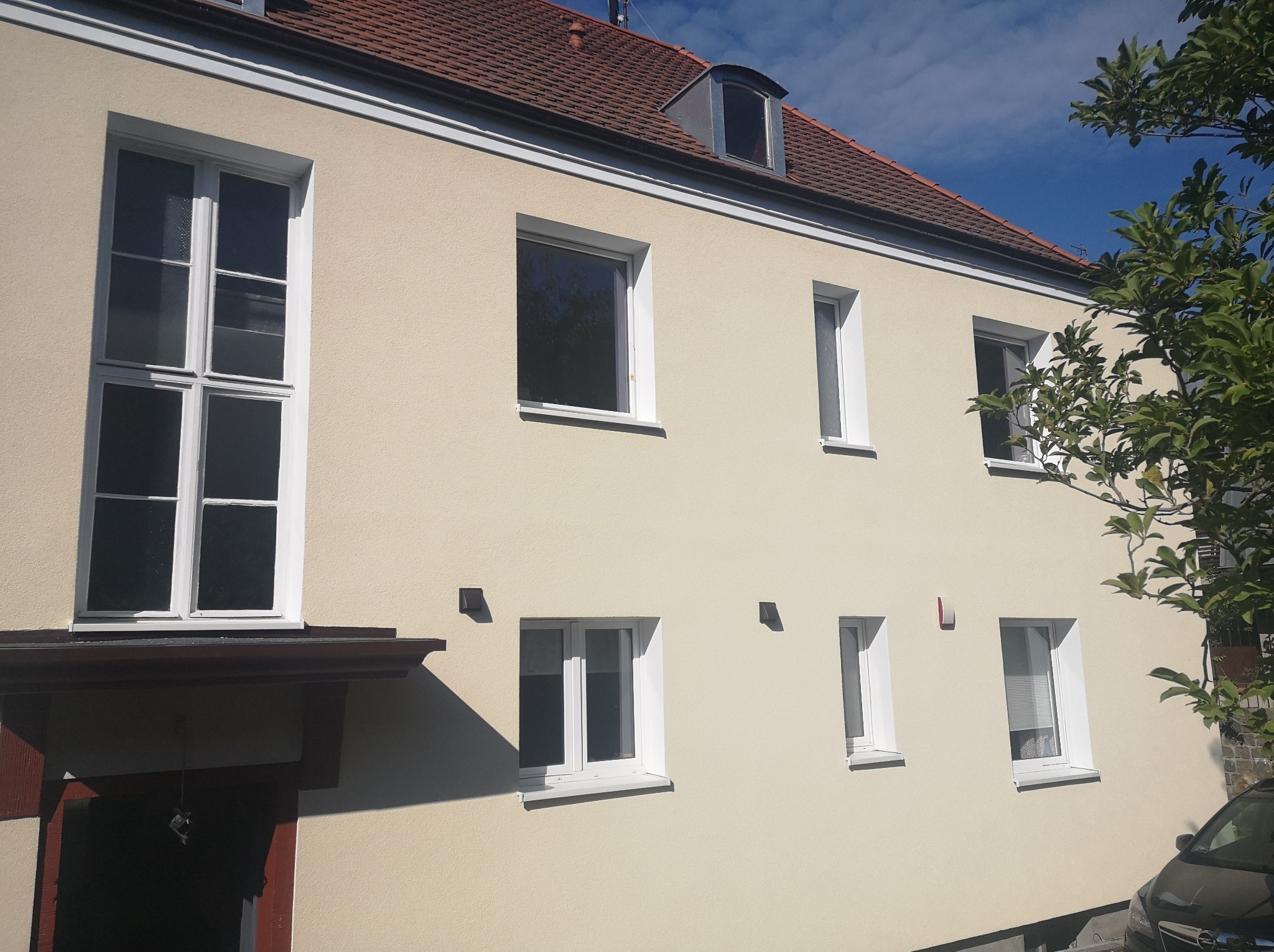 Jasna elewacja budynku wielorodzinnego z oknami o różnej wielkości i kształcie, widoczny fragment dachu z lukarną i zieleni drzew