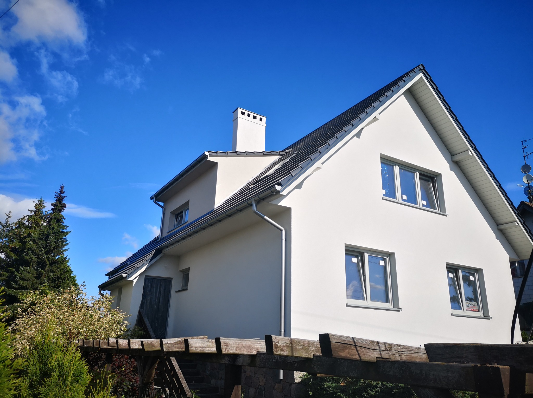 Nowoczesny dom jednorodzinny z białą elewacją, ciemną dachówką i widocznym kominem na tle błękitnego nieba, częściowo zasłonięty drewnianą konstrukcją.