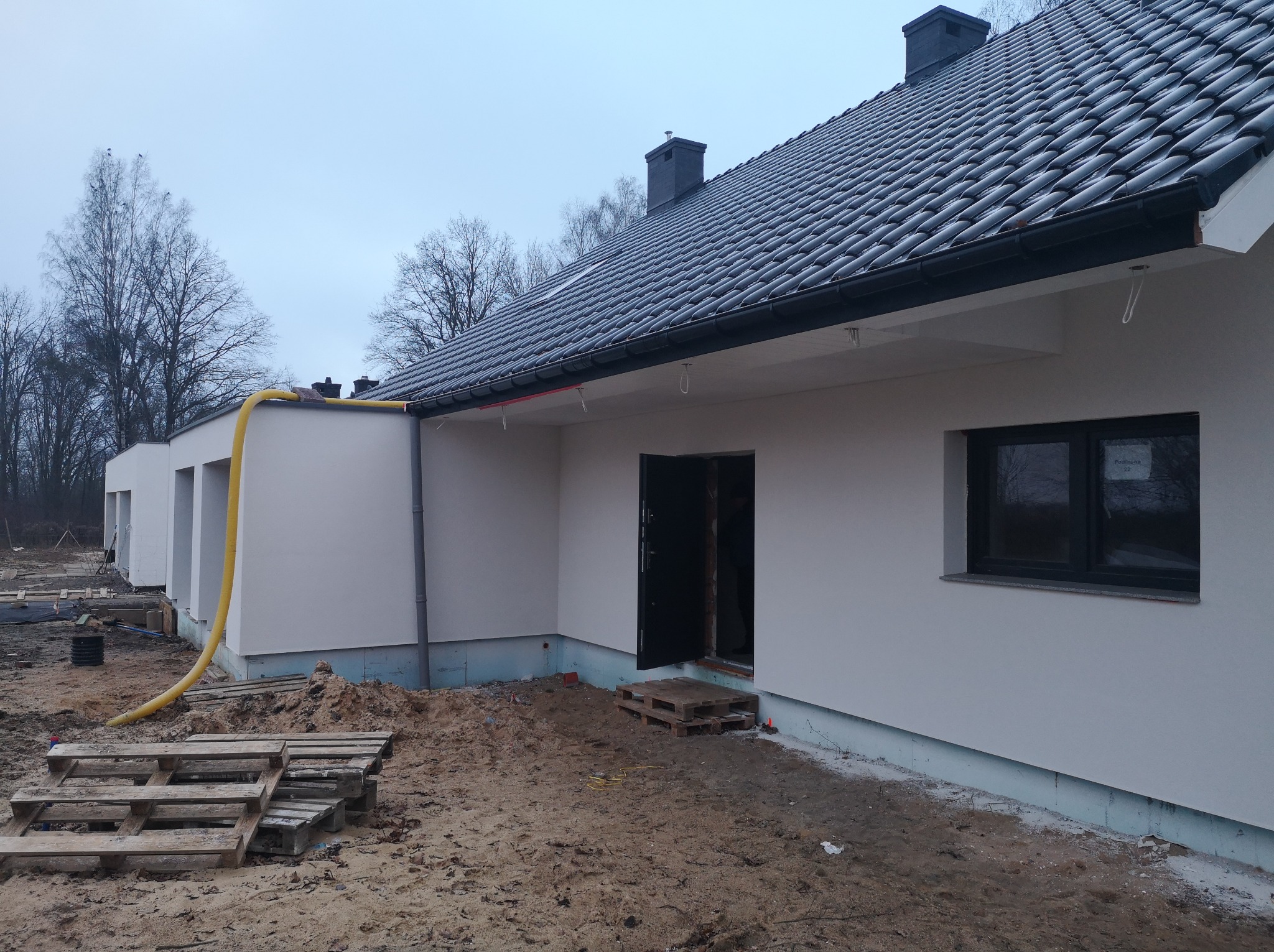 Nowoczesny dom jednorodzinny z ciemną dachówką, jasną elewacją i czarnymi oknami, widoczne palety i żółty wąż na placu budowy, stan surowy zamknięty.