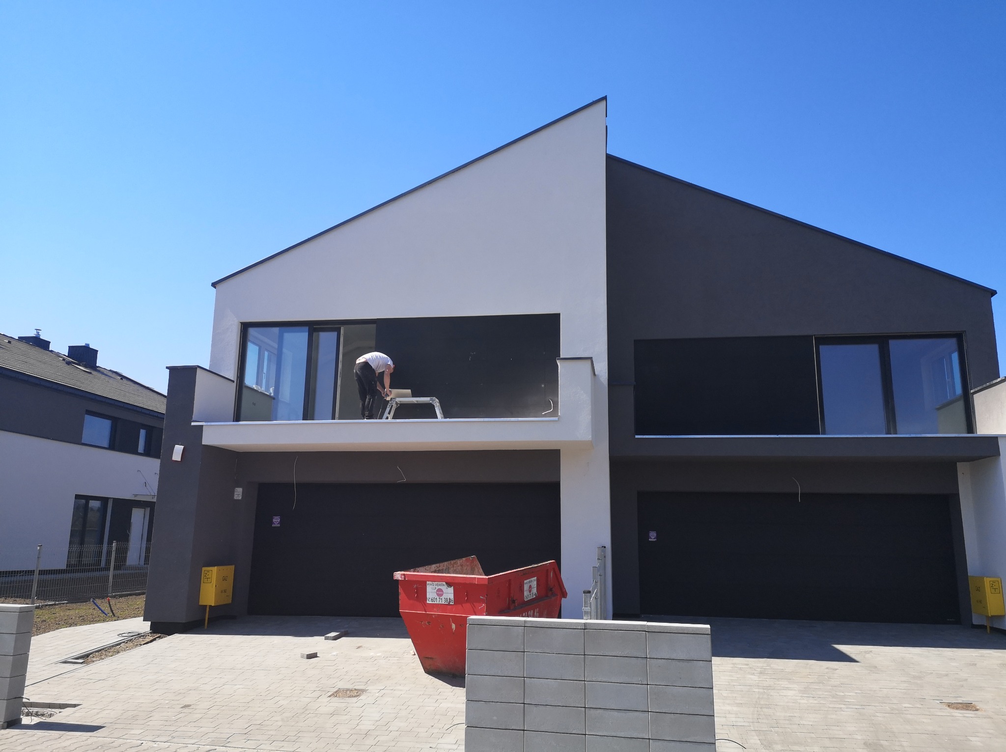 Dwukondygnacyjny dom w trakcie wykańczania elewacji, widoczny pracownik na rusztowaniu na balkonie, czerwony kontener na gruz przed budynkiem, nowoczesna architektura.