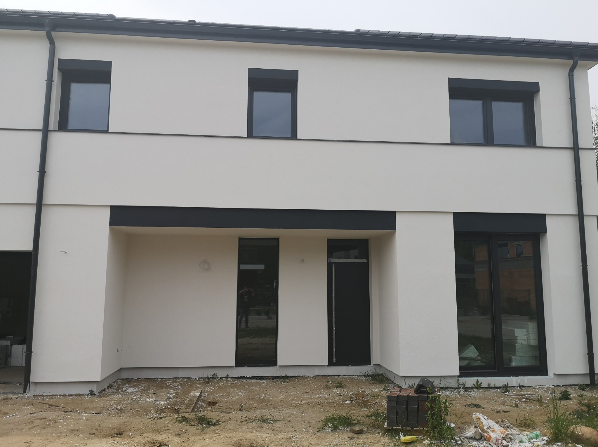 Nowoczesny, dwukondygnacyjny dom z jasną elewacją i czarnymi ramami okiennymi oraz rynnami; widoczne ślady prac wykończeniowych na gruncie.