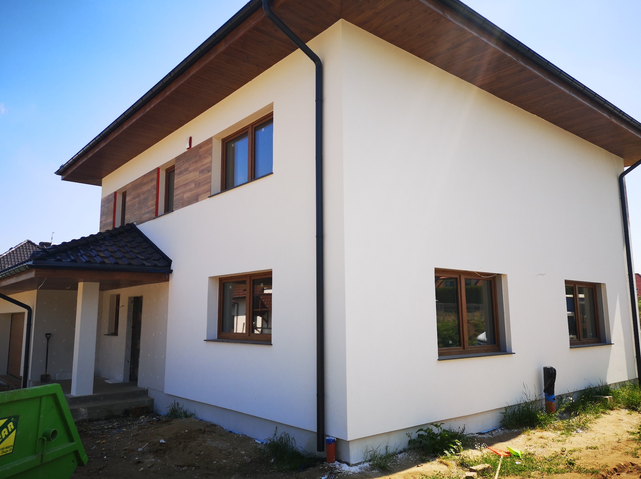 Dwupiętrowy dom z białą elewacją, drewnianymi oknami i podbitką dachową, częściowo wykończony, widoczny kontener na gruz i materiały budowlane.