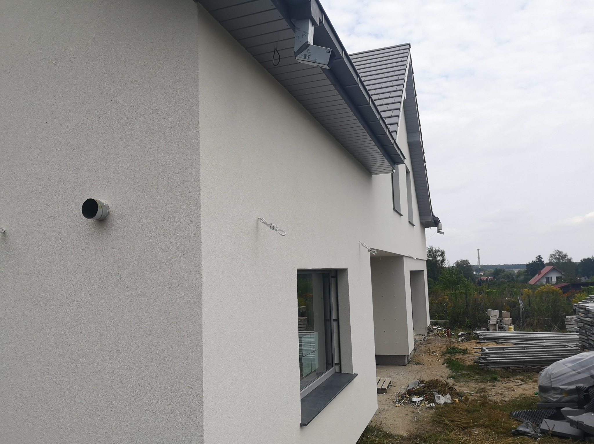 Jasna elewacja domu jednorodzinnego z widocznym systemem rynnowym, okno z ciemnym parapetem i fragmentem terenu budowy z materiałami budowlanymi w tle.