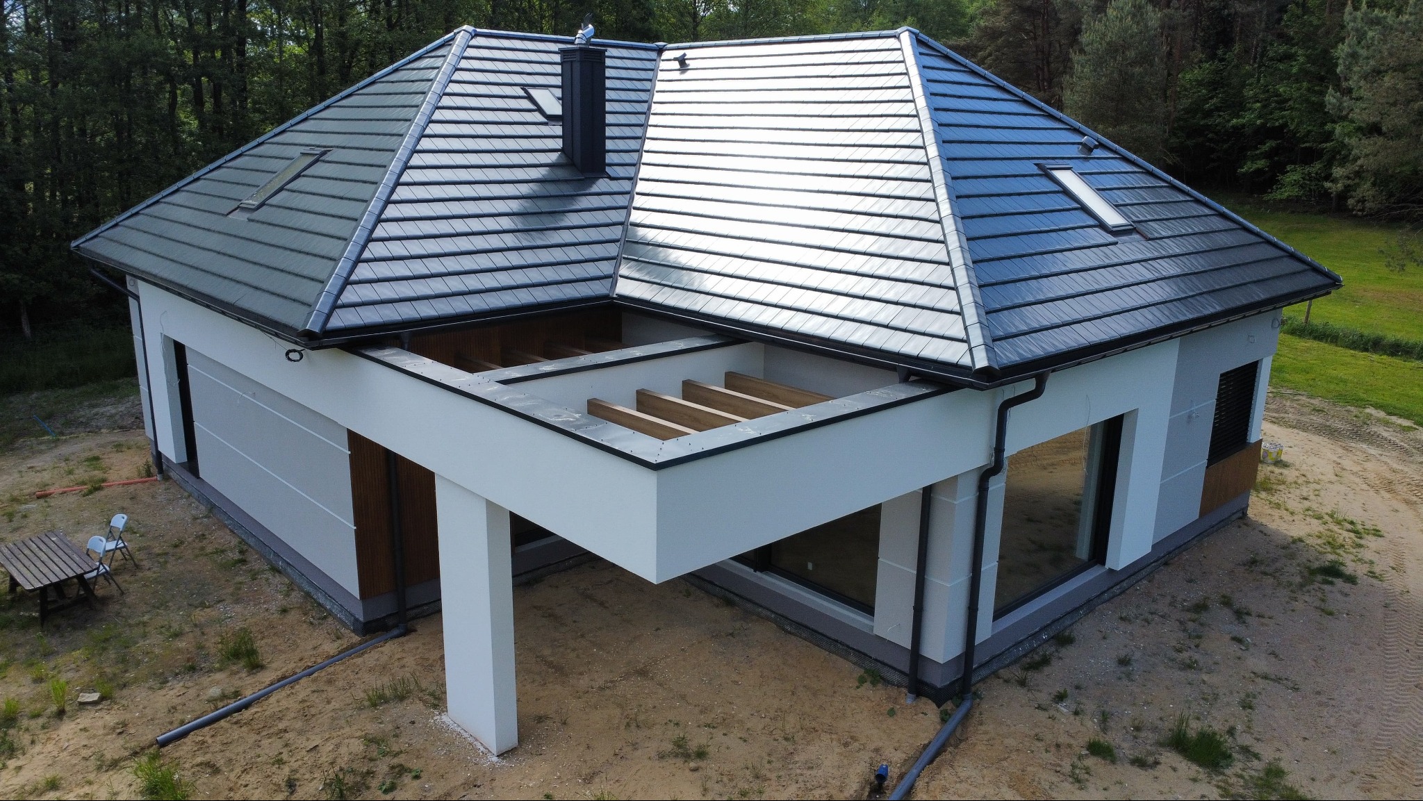 Nowoczesny dom z grafitową dachówką i jasną elewacją, widok z góry. Widoczne elementy konstrukcyjne tarasu i otoczenie w naturalnym krajobrazie.