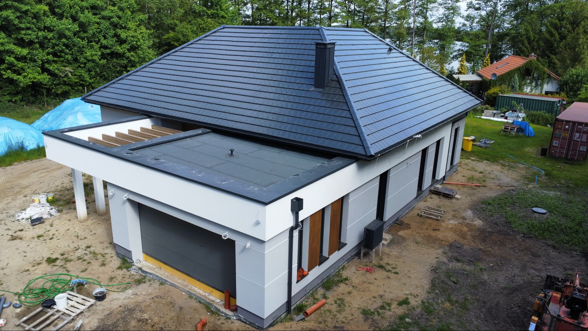 Nowoczesny dom z płaskim dachem i garażem, widok z góry. Szara dachówka i biała elewacja. Elementy wykończeniowe w kolorze antracytowym. Otoczenie w trakcie prac.