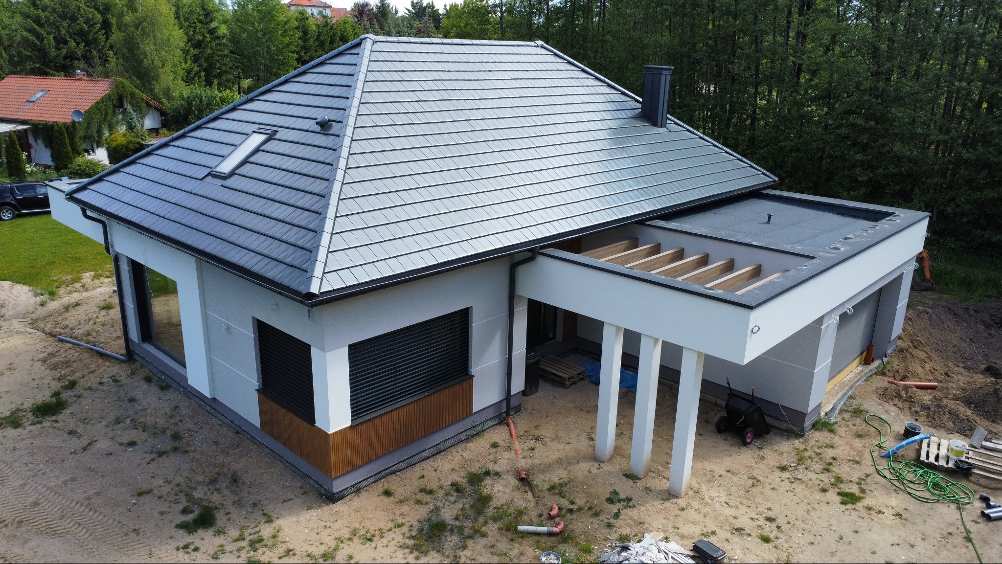 Nowoczesny dom jednorodzinny z szarą dachówką, białymi ścianami i drewnianymi akcentami. Widok z góry ukazuje detale elewacji i otoczenie budynku.