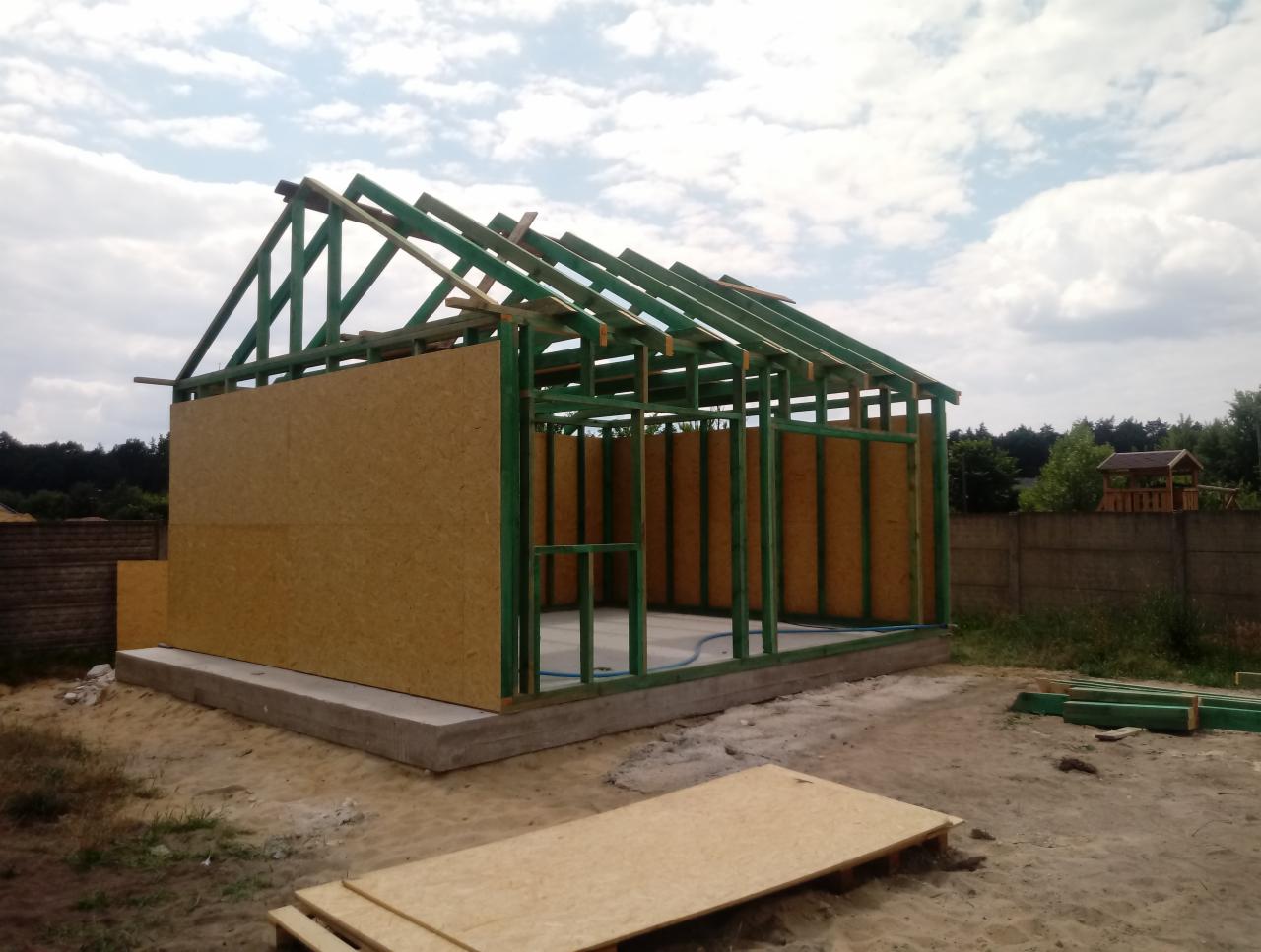 Wznoszenie konstrukcji budynku z drewnianym szkieletem, widoczne płyty OSB, zielone belki, betonowa podstawa, w tle ogrodzenie i altana.