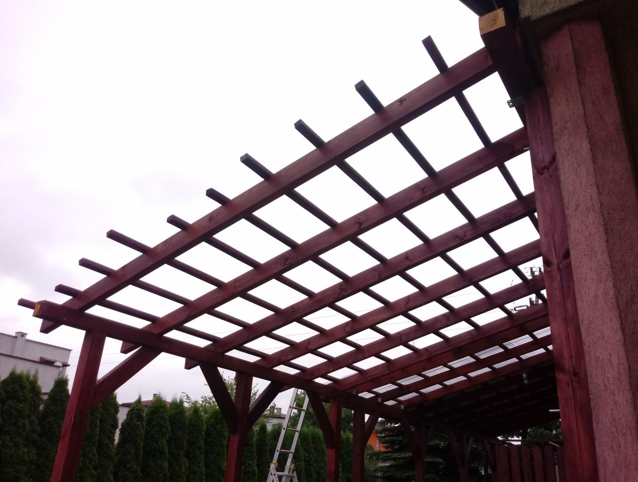 Drewniana konstrukcja z belek, prawdopodobnie altana lub pergola, z widoczną drabiną w tle i zielonymi drzewami.