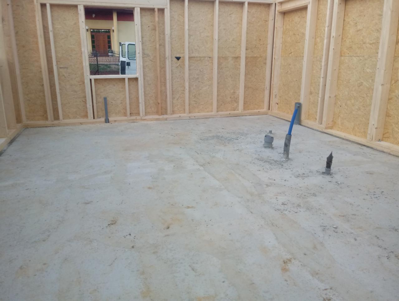 Wnętrze domu szkieletowego w trakcie budowy, widoczne elementy konstrukcyjne ścian z drewna i płyty OSB, surowa betonowa podłoga z wystającymi rurami instalacyjnymi, okno z widokiem na podwórze...