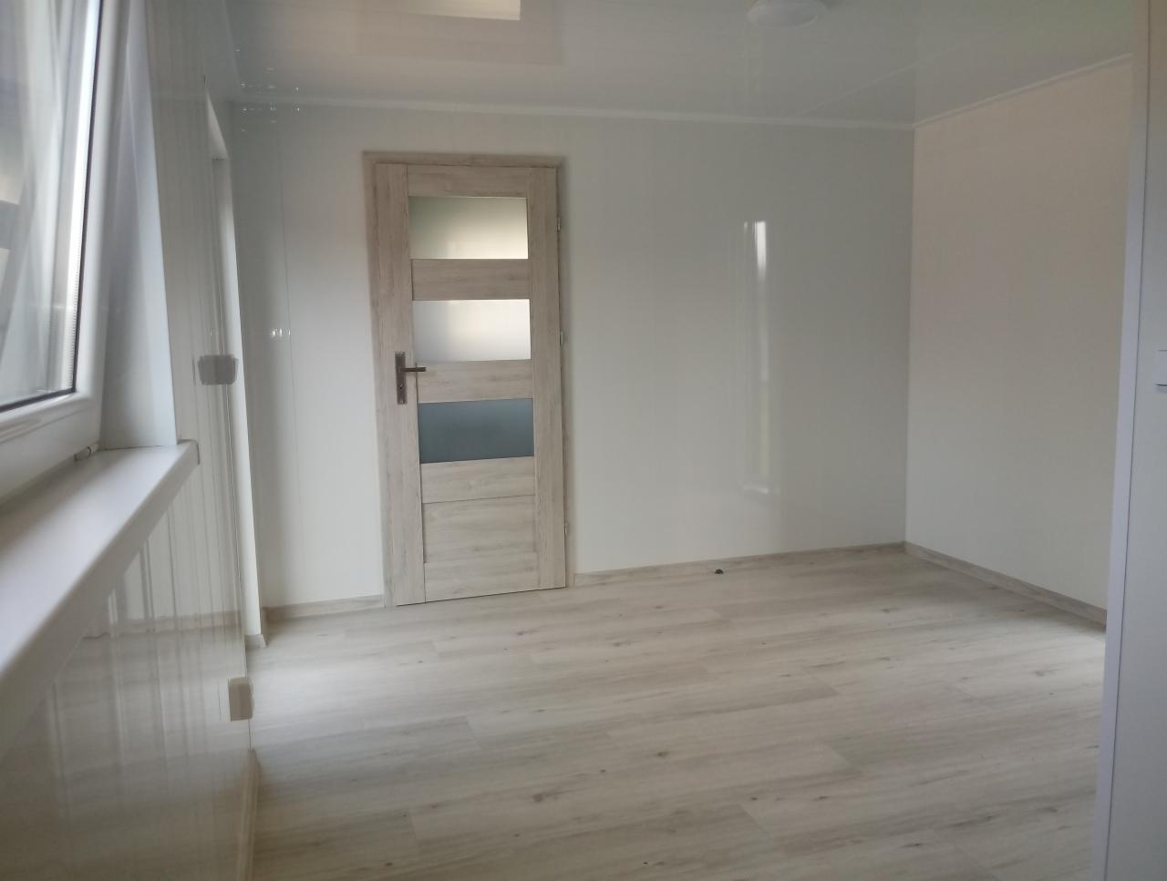 Wnętrze pustego pokoju w domu szkieletowym, z jasnymi ścianami, panelami podłogowymi i drzwiami z przeszkleniami, widok z okna z białymi zasłonami.