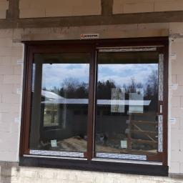 Okna i D-ach! - Brązowe okno z roletą zewnętrzną zamontowane w surowym murze z betonu komórkowego, widoczne odbicie nieba i drzew w szybie, folia ochronna na ramie okiennej.