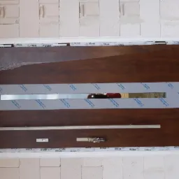 Brązowe drzwi wejściowe z długim, srebrnym uchwytem, zabezpieczone folią ochronną i taśmą, zamontowane w surowym otworze ściany z betonu komórkowego.