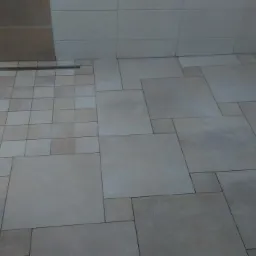 Wykończona łazienka z beżowymi płytkami o zr&oacute;żnicowanych rozmiarach, ułożonych w nieregularny wz&oacute;r, widoczny odpływ liniowy przy ścianie.