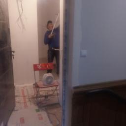 Elpisbud Sp. z o.o - Malowanie ścian wewnątrz mieszkania, widoczne zabezpieczenie podłogi, krzesło z grzejnikiem i malarz z wałkiem w tle.