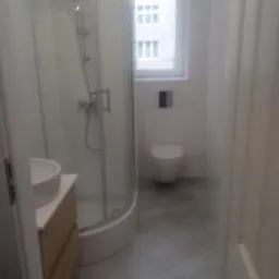 Nowoczesna łazienka z kabiną prysznicową z zaokrąglonym szkłem, umywalką nablatową na szafce z drewnopodobnymi frontami, toaletą podwieszaną i szarymi płytkami podłogowymi w formacie kwadratu.