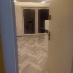 Ułożona podłoga z paneli w jodełkę, widok zza otwartych drzwi, jasne kolory, widoczne drzwi balkonowe.