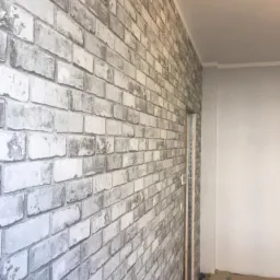 Ściana pokryta szarą tapetą imitującą cegłę, widoczny fragment drzwi i zabezpieczona podłoga.