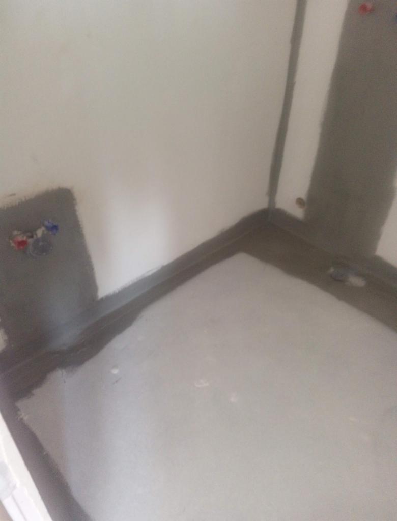 Przygotowanie do prac wykończeniowych w łazience: zabezpieczenie narożników i podłogi szarą masą uszczelniającą wokół rur doprowadzających wodę, widoczny odpływ podłogowy.