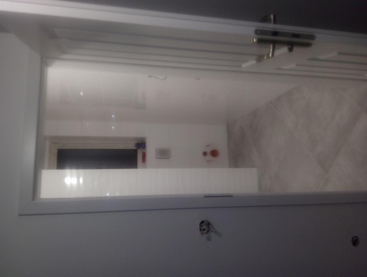 Widok na wykończoną łazienkę przez otwarte drzwi, z widocznymi białymi płytkami na ścianach, szarymi płytkami podłogowymi ułożonymi w romby oraz czarnym oknem w tle.