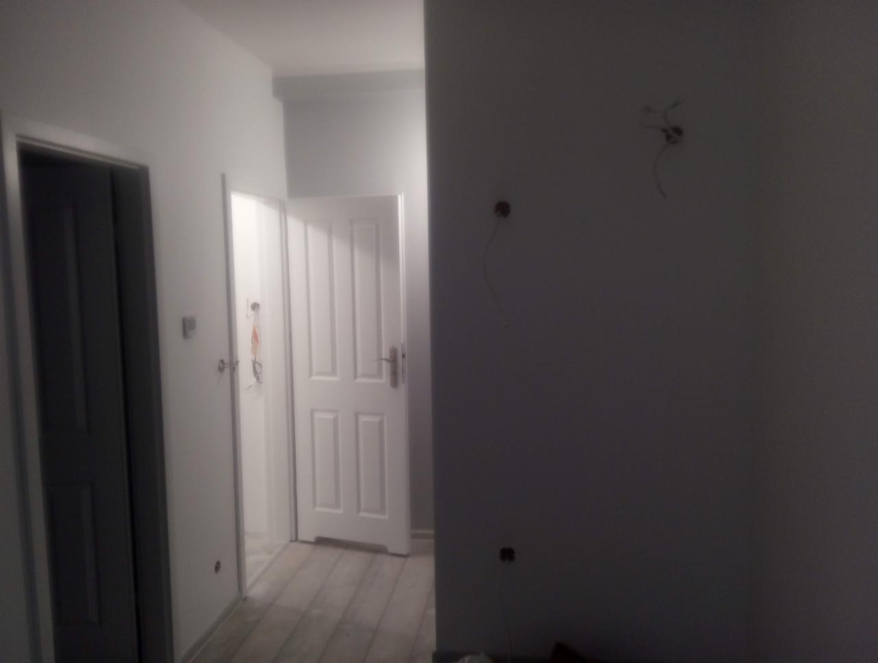 Korytarz w trakcie odświeżania: widoczne dwie białe, częściowo otwarte drzwi po lewej stronie, szare panele podłogowe, ściany przygotowane do malowania z wystającymi kablami.