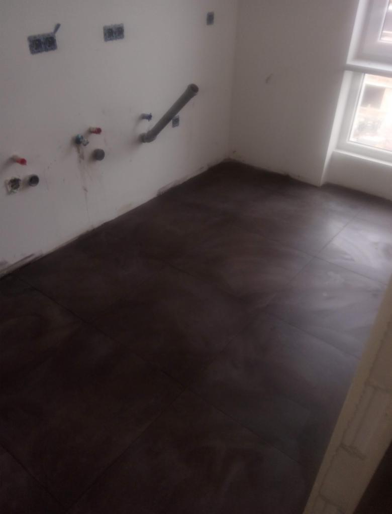 Ułożone ciemnobrązowe płytki podłogowe w pomieszczeniu w trakcie remontu, widoczne instalacje hydrauliczne na ścianie i okno w tle.