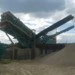 Duża, zielona maszyna do przesiewania kruszywa na placu budowy, z widocznymi taśmociągami i hałdą piasku.