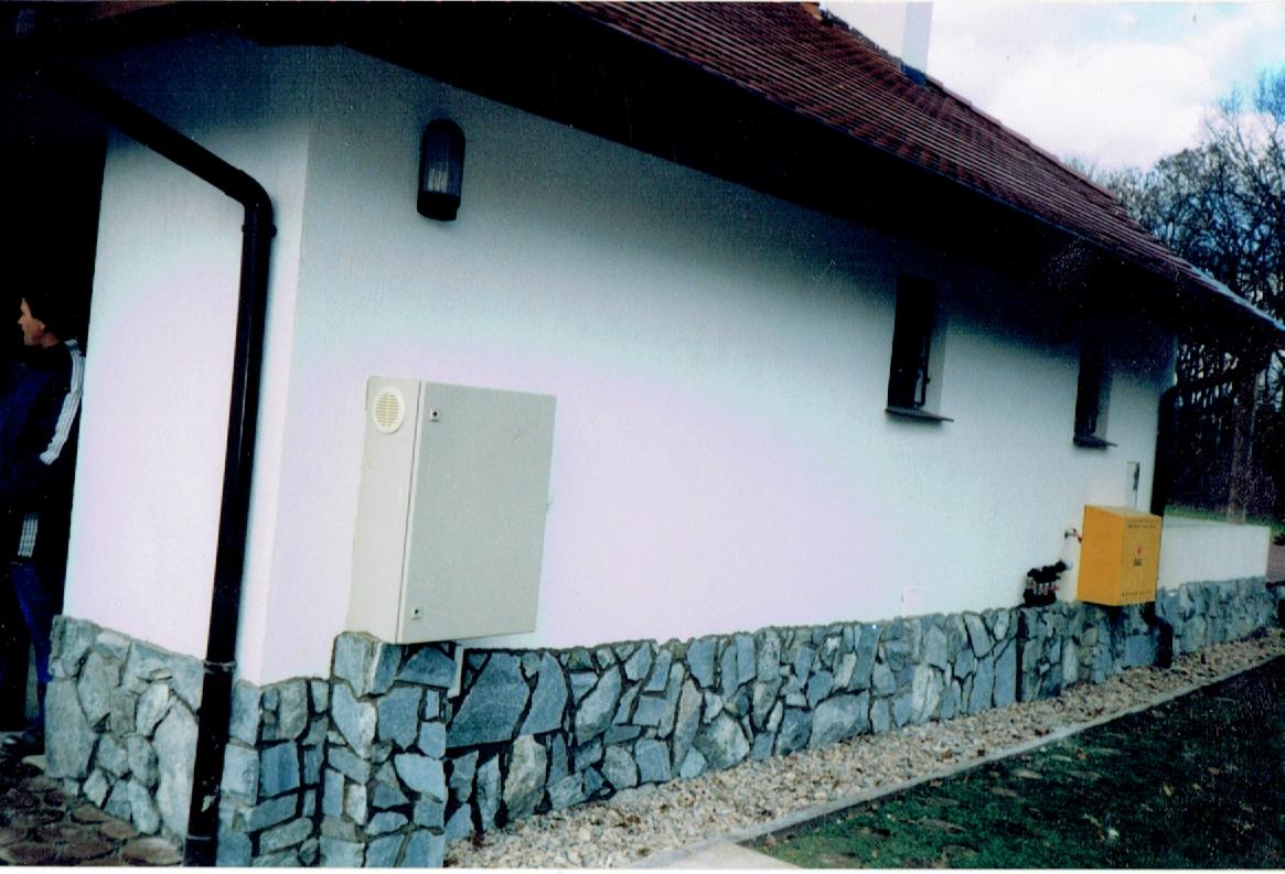 Boczny widok domu z białą elewacją, kamienną podmurówką i brązową dachówką, widoczna skrzynka elektryczna na ścianie i fragment osoby w tle.