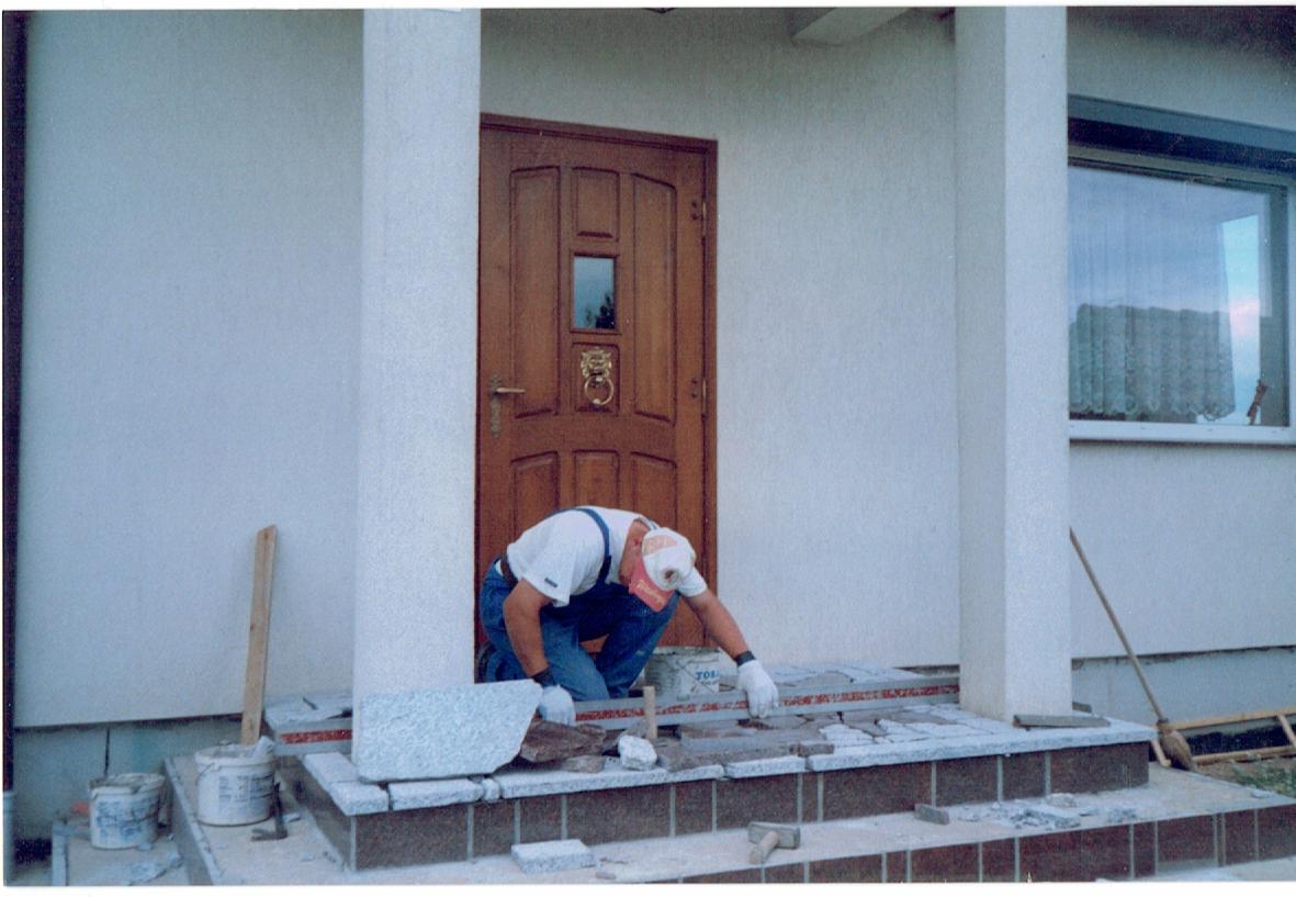 Pracownik w białej koszulce i niebieskich spodniach, z czerwoną czapką na głowie, układa granitowe płytki na schodach wejściowych do domu z brązowymi drzwiami i kolumnami, używając poziomicy i młotka.