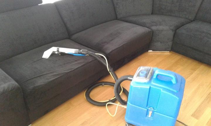 Czyszczenie ciemnej, narożnej kanapy za pomocą niebieskiego odkurzacza piorącego z białą końcówką na drewnianej podłodze.