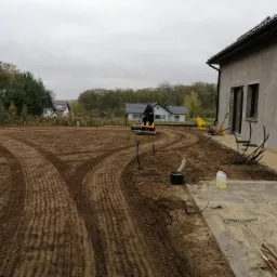 Przygotowanie terenu pod trawnik: operator na glebogryzarce wyrównuje glebę na działce przy nowoczesnym domu, widoczne ślady opon i resztki ziemi na kostce.