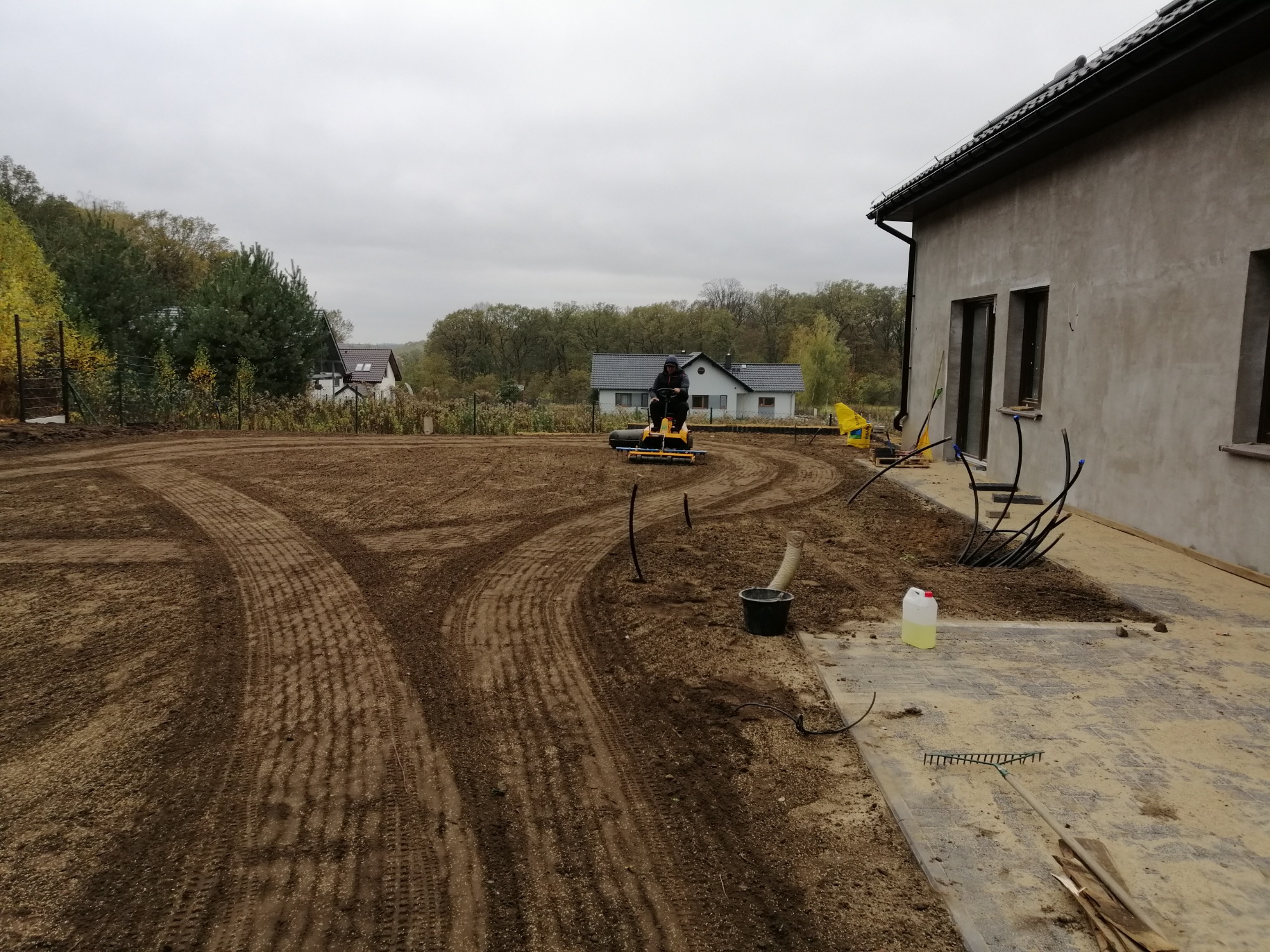 Przygotowanie terenu pod trawnik: operator na glebogryzarce wyrównuje glebę na działce przy nowoczesnym domu, widoczne ślady opon i resztki ziemi na kostce.