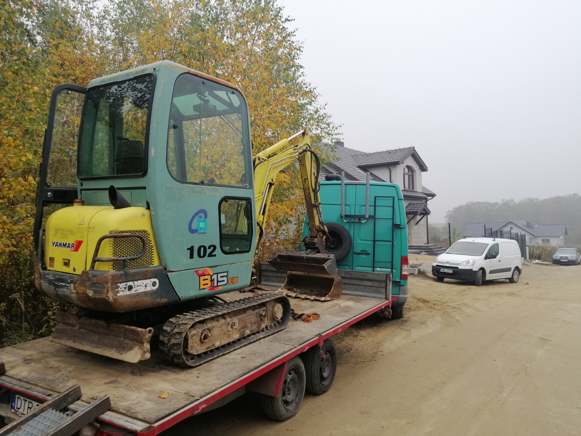 Niewielka, używana koparka Yanmar, oznaczona numerem 102, transportowana na lawecie, w tle zielony bus i zabudowa domów jednorodzinnych w pochmurny dzień.
