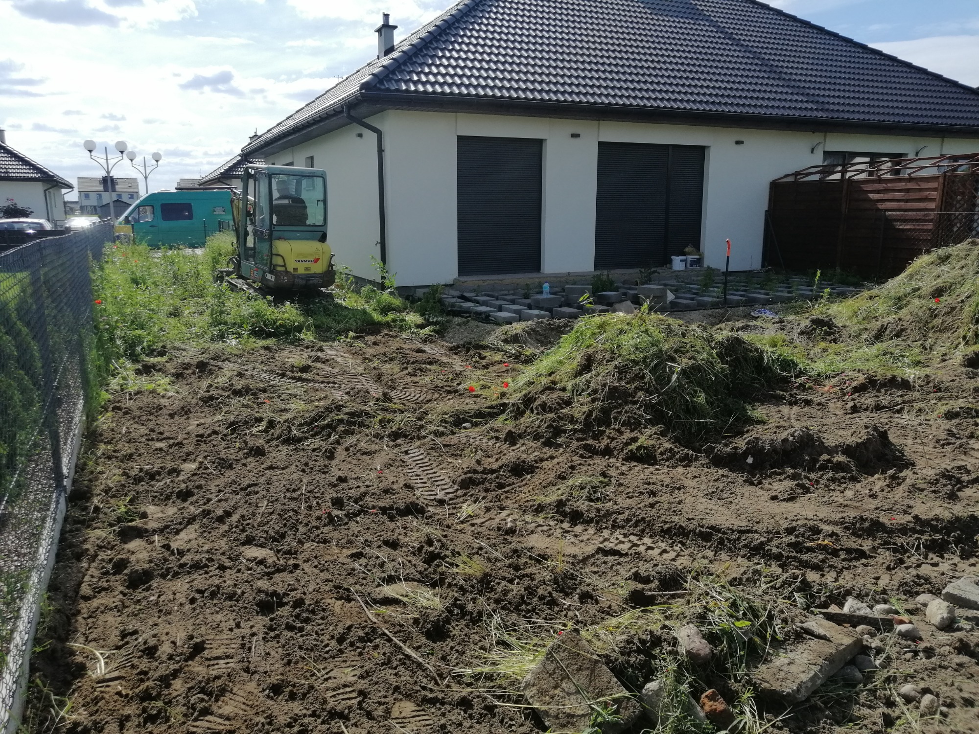 Mała koparka na działce z przygotowanym terenem, widoczne ślady opon, usunięta darń w pryzmach, w tle budynek z dwoma garażami.