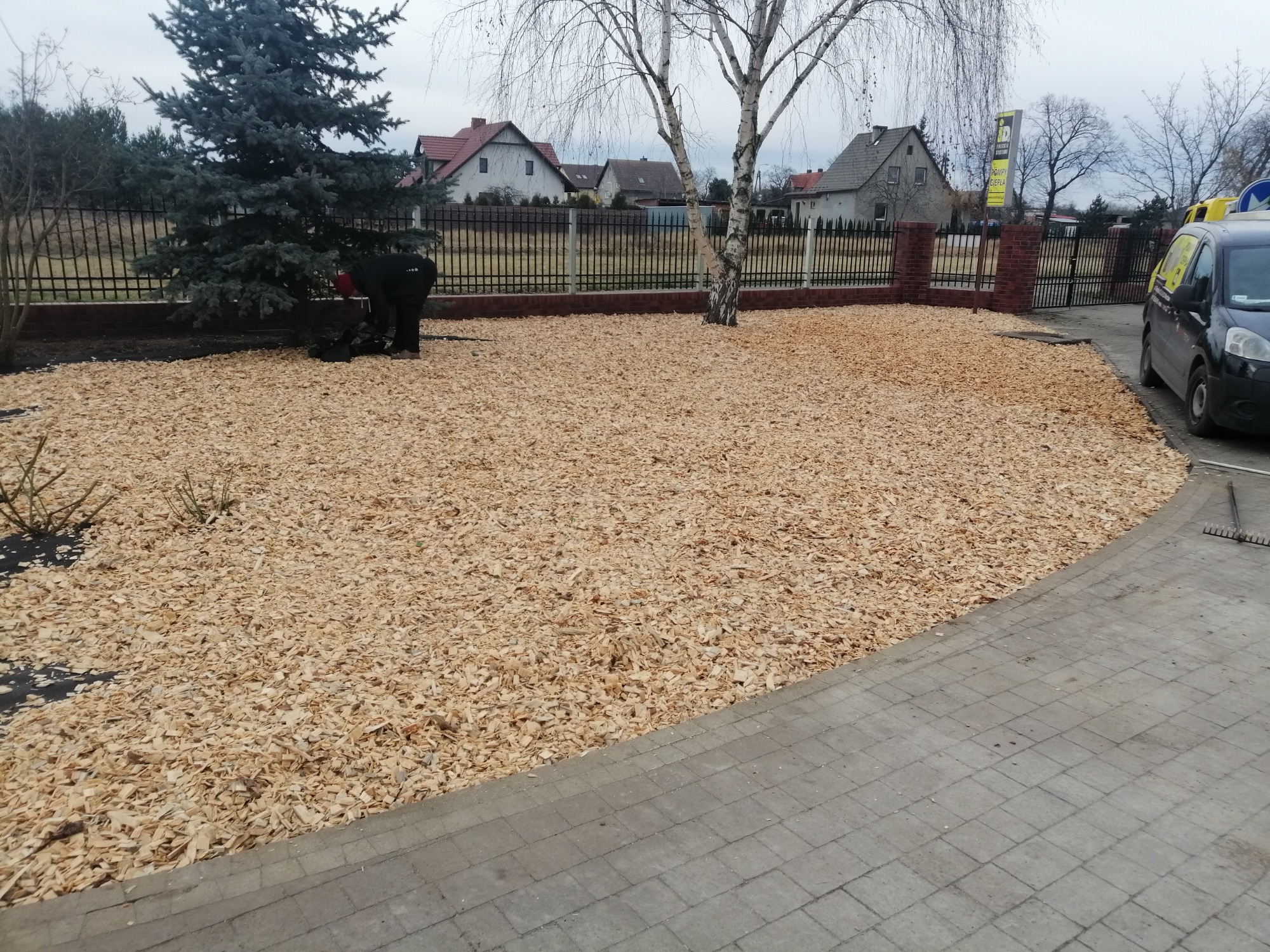 Obszar pokryty świeżymi zrębkami drewna, z widoczną osobą pracującą przy zielonym drzewie iglastym, ogrodzeniem posesji i domami w tle, oraz zaparkowanym samochodem dostawczym.